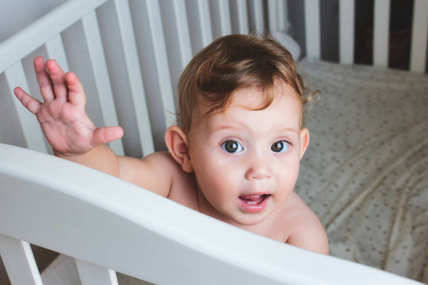 niedliches Baby mit lockigem blondem Haar, das in einem Kinderbett steht, in die Kamera blickt und winkt - Foto, Bild