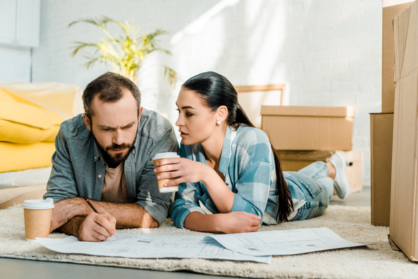 handsome husband and wife lying on floor, drinking coffee and working on blueprints of new house at home, moving concept - Zdjęcie, obraz