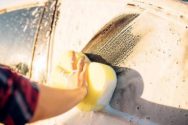 Γυναικείο πρόσωπο χέρι με σφουγγάρι τριψίματος οχήματος με αφρό, πλύσιμο αυτοκινήτων. Νεαρή γυναίκα στο self service αυτοκίνητο πλύσιμο. Εξωτερική carwash - Φωτογραφία, εικόνα