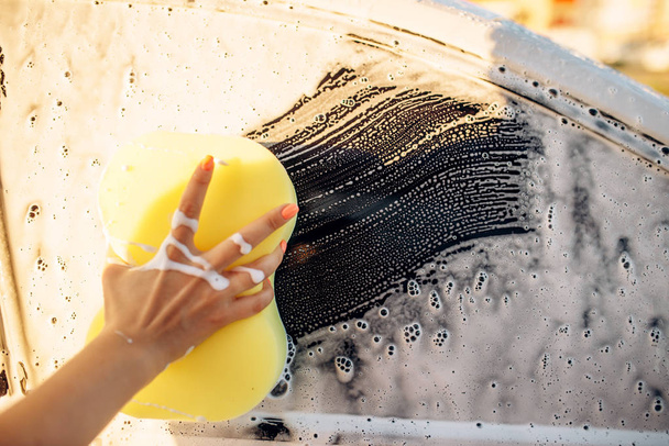Γυναικείο πρόσωπο χέρι με σφουγγάρι τριψίματος οχήματος με αφρό, πλύσιμο αυτοκινήτων. Νεαρή γυναίκα στο self service αυτοκίνητο πλύσιμο. Εξωτερική carwash στη θερινή ημέρα  - Φωτογραφία, εικόνα