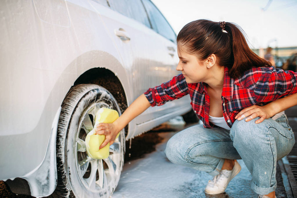 Cute woman with sponge scrubbing vehicle wheel with foam, car wash. Lady on self-service automobile washing. Outdoor carwash at summer day  - Фото, изображение