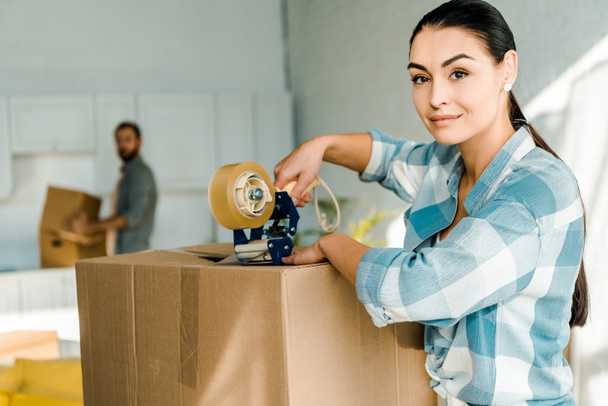 wife packing cardboard box with scotch tape and husband behind, moving concept - Photo, Image