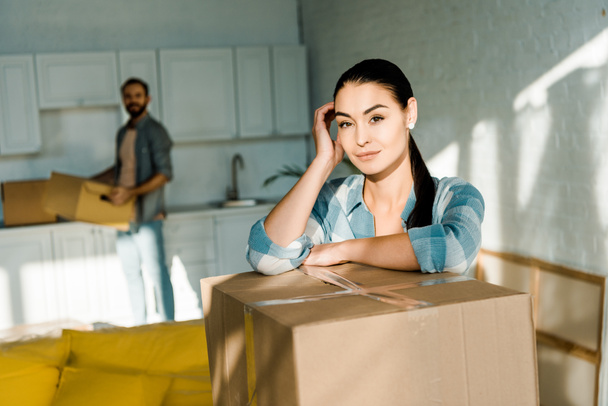 wife leaning on cardboard box with husband packing for new house on background, moving concept - Photo, Image