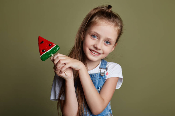 Beautiful young smiling teen girl holding a Lollipop in the form of a slice of watermelon. Cheerful joyful emotions on the girl face - Zdjęcie, obraz
