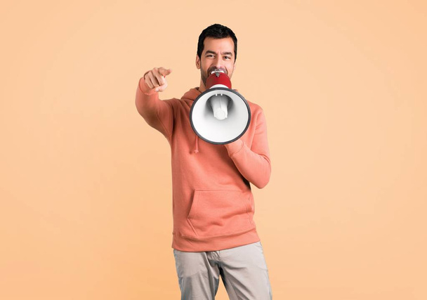 Man in a pink sweatshirt shouting through a megaphone to announce something while pointing to the front on ocher background - Фото, зображення