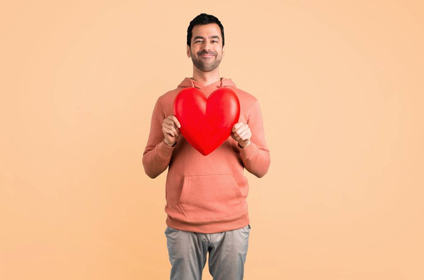 Man in a pink sweatshirt holding a big heart icon toy on ocher background - Foto, Imagem