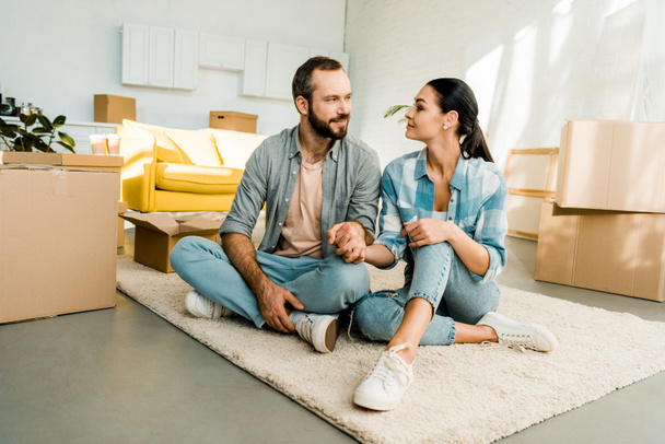husband and wife sitting on floor and holding hands while packing for new house, moving concept - Photo, Image