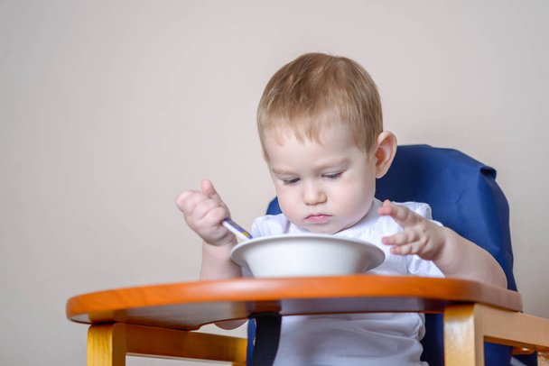 Маленький мальчик учится есть за столом для больших детей, изучает тарелку и ложку на кухне
 - Фото, изображение