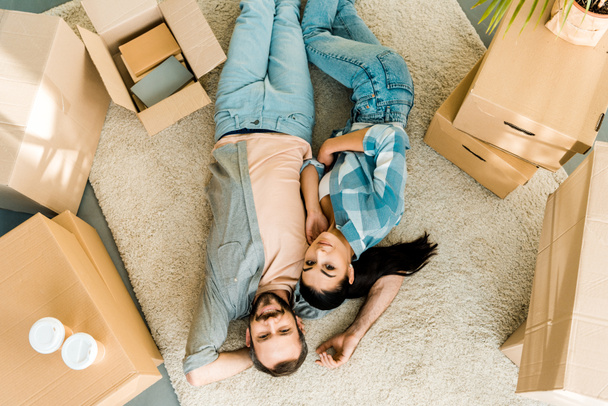 Blick von oben auf lächelndes Paar, das auf dem Boden liegt und sich nach dem Packen für das neue Haus entspannt, bewegtes Konzept - Foto, Bild