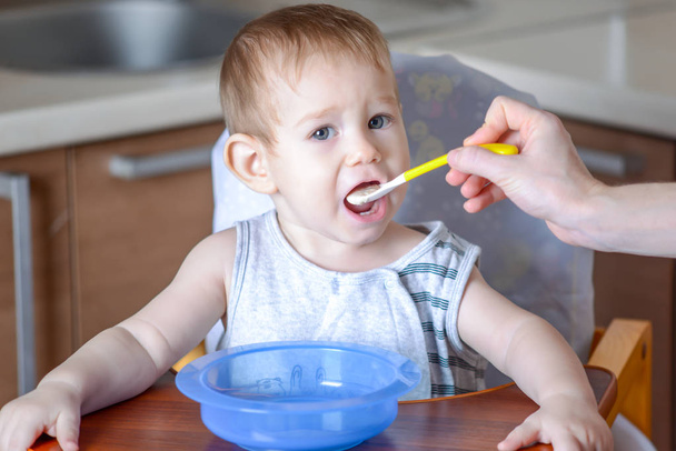 Der kleine Junge isst mit weit aufgerissenem Mund auf einem Stuhl in der Küche sitzend. Mama füttert mit einem Löffel Brei in der Hand - Foto, Bild