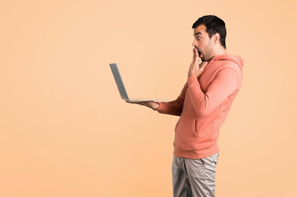 Surprised man in a pink sweatshirt with laptop on ocher background - Фото, изображение