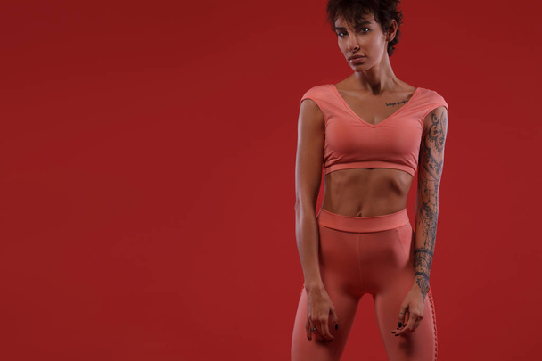Fitness and sport motivation.. Strong and fit athletic, woman with tattoo posing on red background in sportswear. - Photo, Image