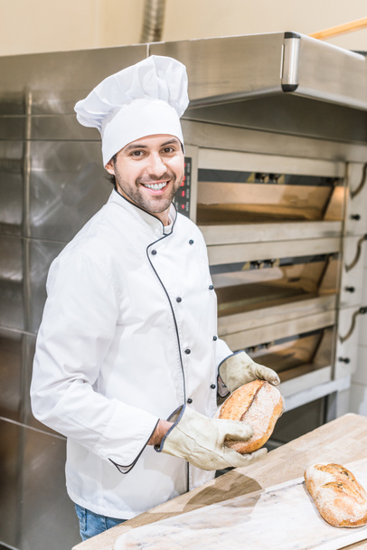 smiling baker in chefs uniform holding fresh bread near oven - Photo, Image