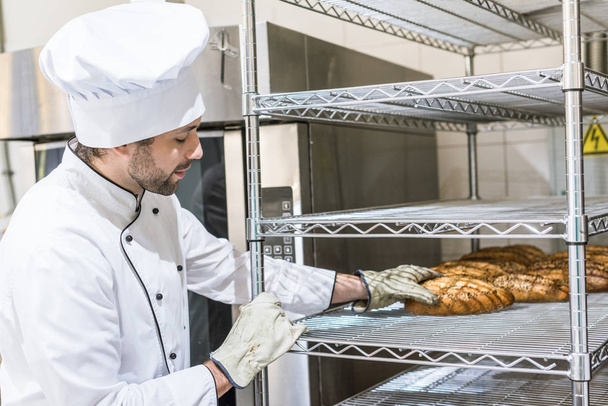 volwassen mannelijke baker aanraken van vers gebakken brood op keuken rek - Foto, afbeelding