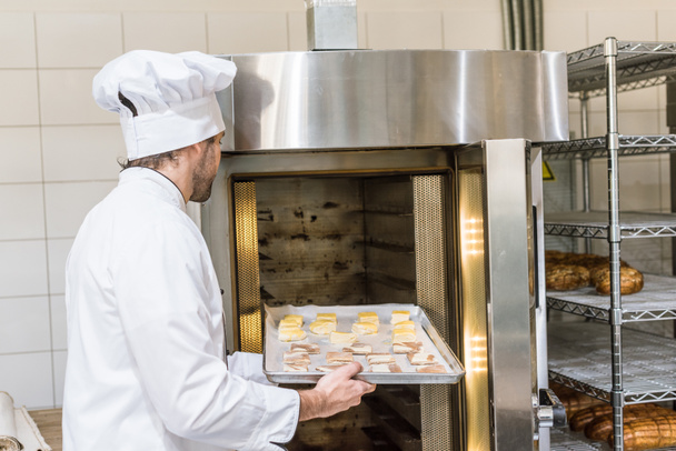 male baker in chefs uniform putting raw dough in oven - Foto, imagen