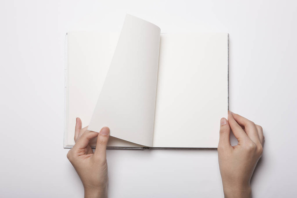 Το γυναικείο χέρι κρατά ένα άδειο βιβλίο απλωμένο στο λευκό τραπέζι - Φωτογραφία, εικόνα