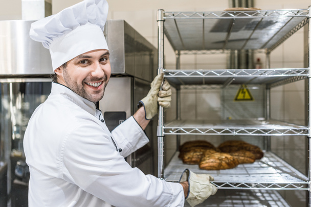 panadero masculino sonriente con pan horneado caliente en rack
 - Foto, Imagen