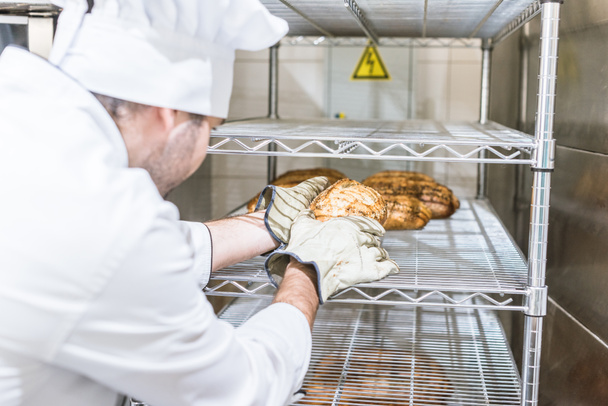 male baker in white chefs uniform putting fresh hot bread at rack  - Foto, afbeelding
