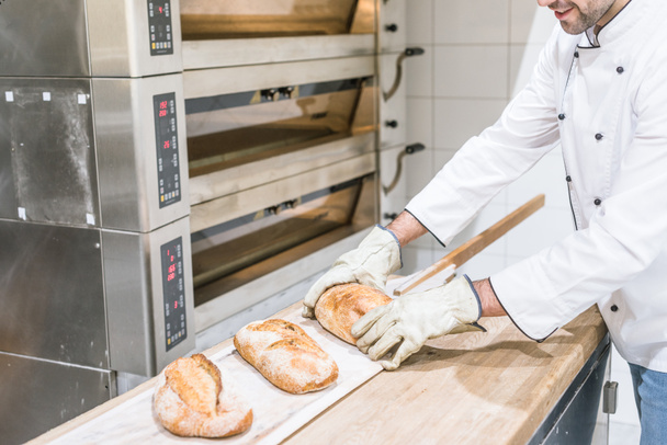 panadero de pie cerca del horno con pan caliente horneado en el mostrador de madera
 - Foto, Imagen