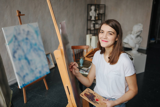 Boldog mosolygó művész festő fiatal gyönyörű lány. Munka folyamat létrehozása. festmény a festőállvány. ihletett munkája. Vízszintes összetétele - Fotó, kép