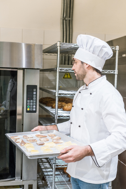 Erwachsener Bäcker in weißer Kochuniform legt Backblech mit ungekochtem Teig in den Ofen - Foto, Bild