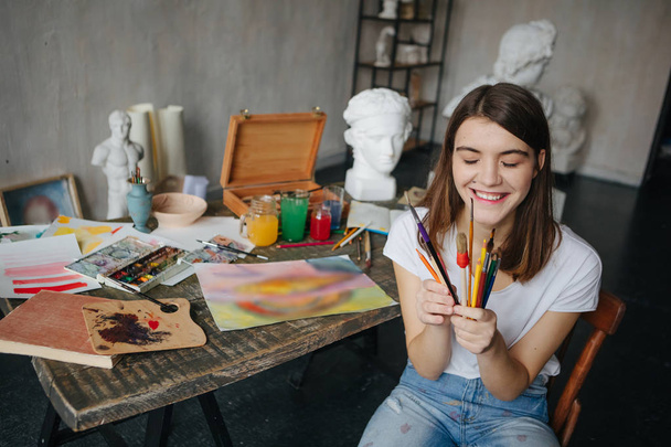 Adorable young artist girl holding brushes and smiling. Creative workshop room at the background. Happy moments - Photo, image