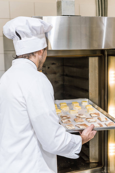 male baker in white chefs uniform putting baking tray with uncooked dough in oven - Fotó, kép