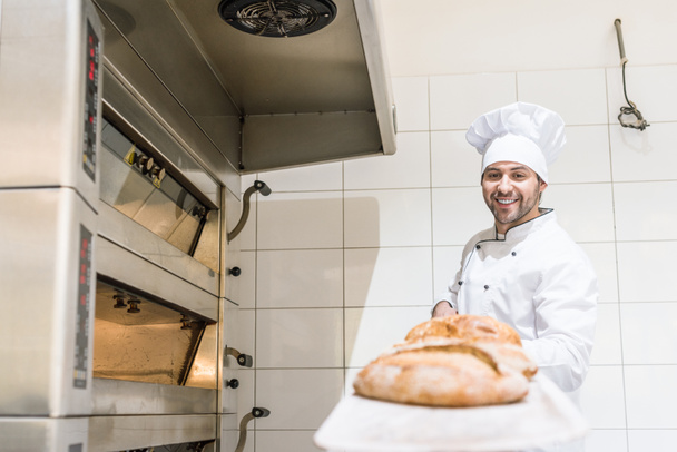 smiling baker in white chefs hat holding board with hot fresh bread - Photo, Image
