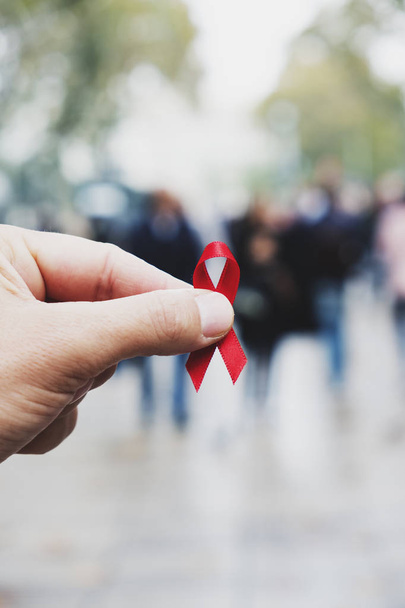 close-up de uma fita de consciência vermelha para a luta contra a AIDS na mão de um jovem caucasiano em uma rua pedonal movimentada de uma cidade
 - Foto, Imagem