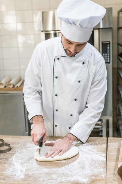 Panadero en chefs blancos uniforme de corte de masa en la mesa de madera en la cocina profesional
 - Foto, Imagen