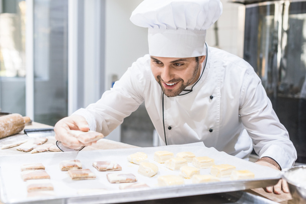 Panadero en uniforme de chefs sonriendo y poniendo masa sin cocer en bandeja
 - Foto, Imagen