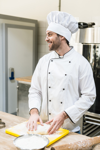 Cuisiner en chef uniforme souriant et pétrissant pâte sur planche à découper
 - Photo, image
