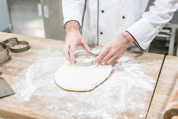 Vista recortada de manos de panadero cortando masa con cortador redondo en mesa de madera
 - Foto, Imagen