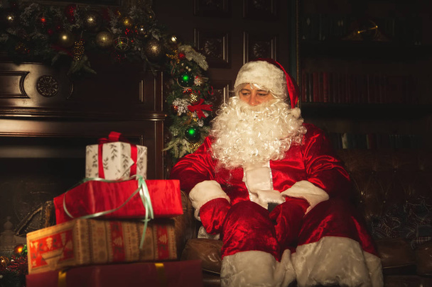 Authentic Santa Claus is sitting in the living room near the Christmas tree. The atmosphere of celebration and magic. - Foto, imagen