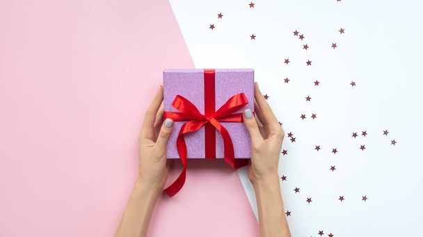 Woman hands holding gift with bow on pink and white background with copy space. Flat lay. - 写真・画像