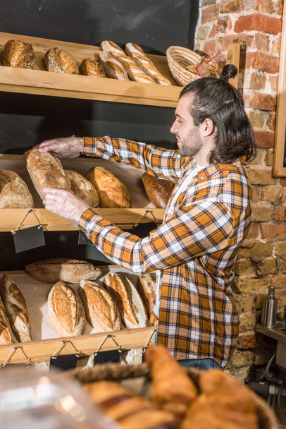 Side view of male seller putting freshly baked bread on wooden shelf  - Foto, imagen