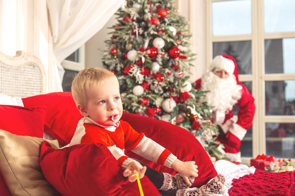 Little sad boy is sitting on a bed in a festive bedroom. The real Santa Claus is hiding from him behind the Christmas tree. - Фото, зображення