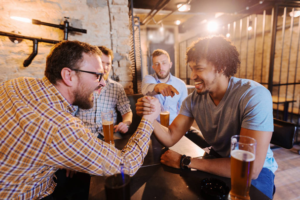 young happy men drinking with beer in pub and having arm wrestling challenge  - Photo, Image