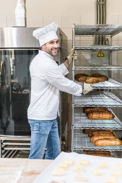 Zijaanzicht van lachende chef nemend rack met brood - Foto, afbeelding