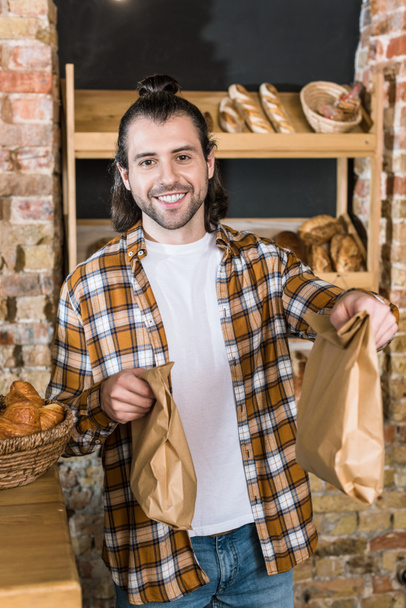 Smiling male seller holding paper bags in bakehouse - Foto, afbeelding