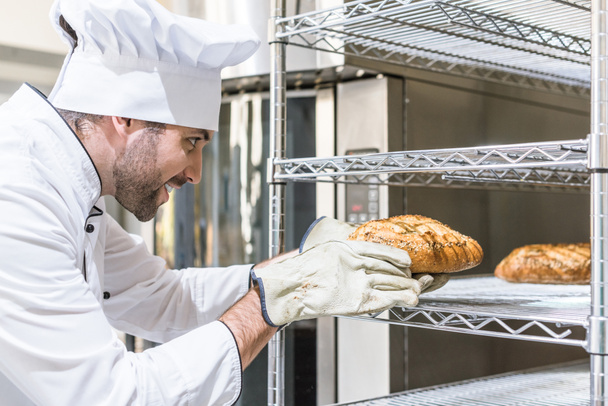 Widok z boku szefa kuchni biorąc świeżo pieczony chleb z półki  - Zdjęcie, obraz