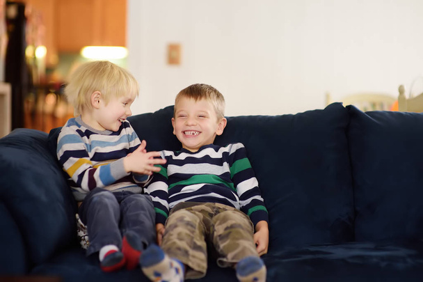 Dwa cute chłopców w paski bluzy, rozmawiając i śmiejąc się, siedząc na kanapie. Pojęcie przyjaźni dziecko. - Zdjęcie, obraz