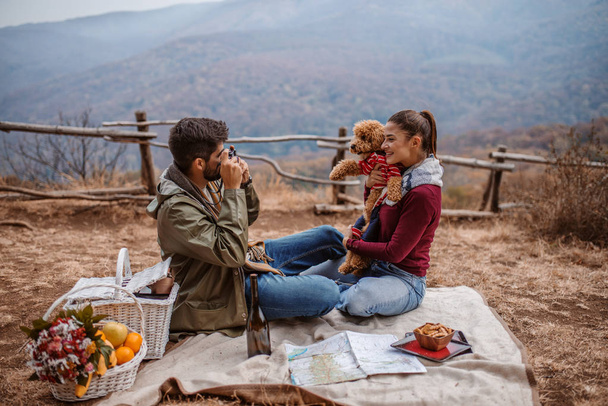 Couple sitting on blanket at picnic. Man taking photo of his girlfriend and dog. Next to them picnic basket and fruit basket. Autumn time, in background mountains. - Foto, imagen