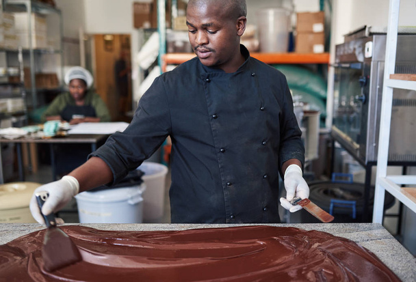 Worker using spatulas to spread melting chocolate on a table while working in an artisanal chocolate making factory - Фото, изображение