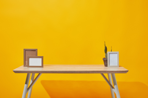 Holztisch mit leeren Bilderrahmen und Kaktus im Blumentopf auf gelb - Foto, Bild