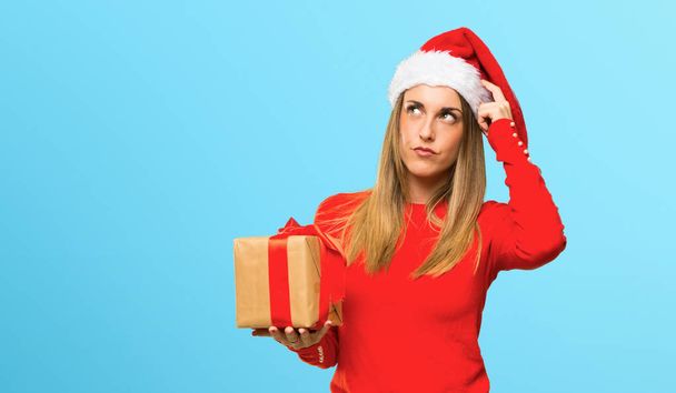 Mulher loira vestida para feriados de Natal com dúvidas e com rosto confuso enquanto coça a cabeça no fundo azul
 - Foto, Imagem