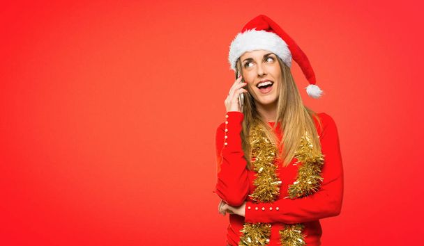Blond žena oblečená vánoční svátky udržet konverzaci s mobilním telefonem s někým na červeném pozadí - Fotografie, Obrázek