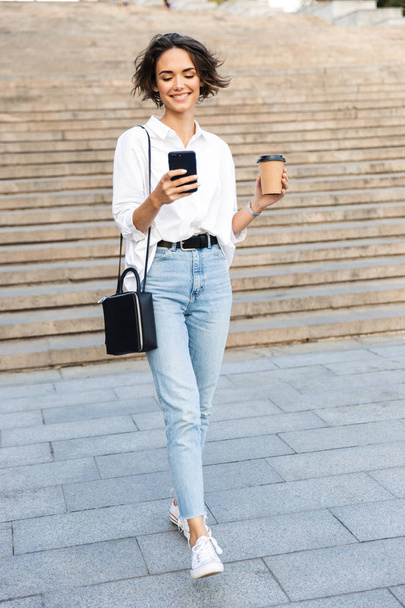 Beautiful young woman walking downstairs at the street, talking on mobile phone, drinking coffee - 写真・画像