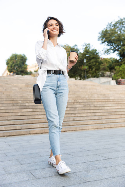 Attractive young woman walking downstairs at the street, talking on mobile phone, drinking coffee - Photo, Image