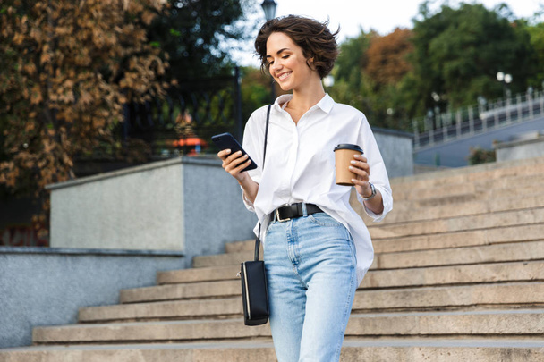 Photo of young cute beautiful woman walking on the street using mobile phone holding coffee. - Foto, Bild